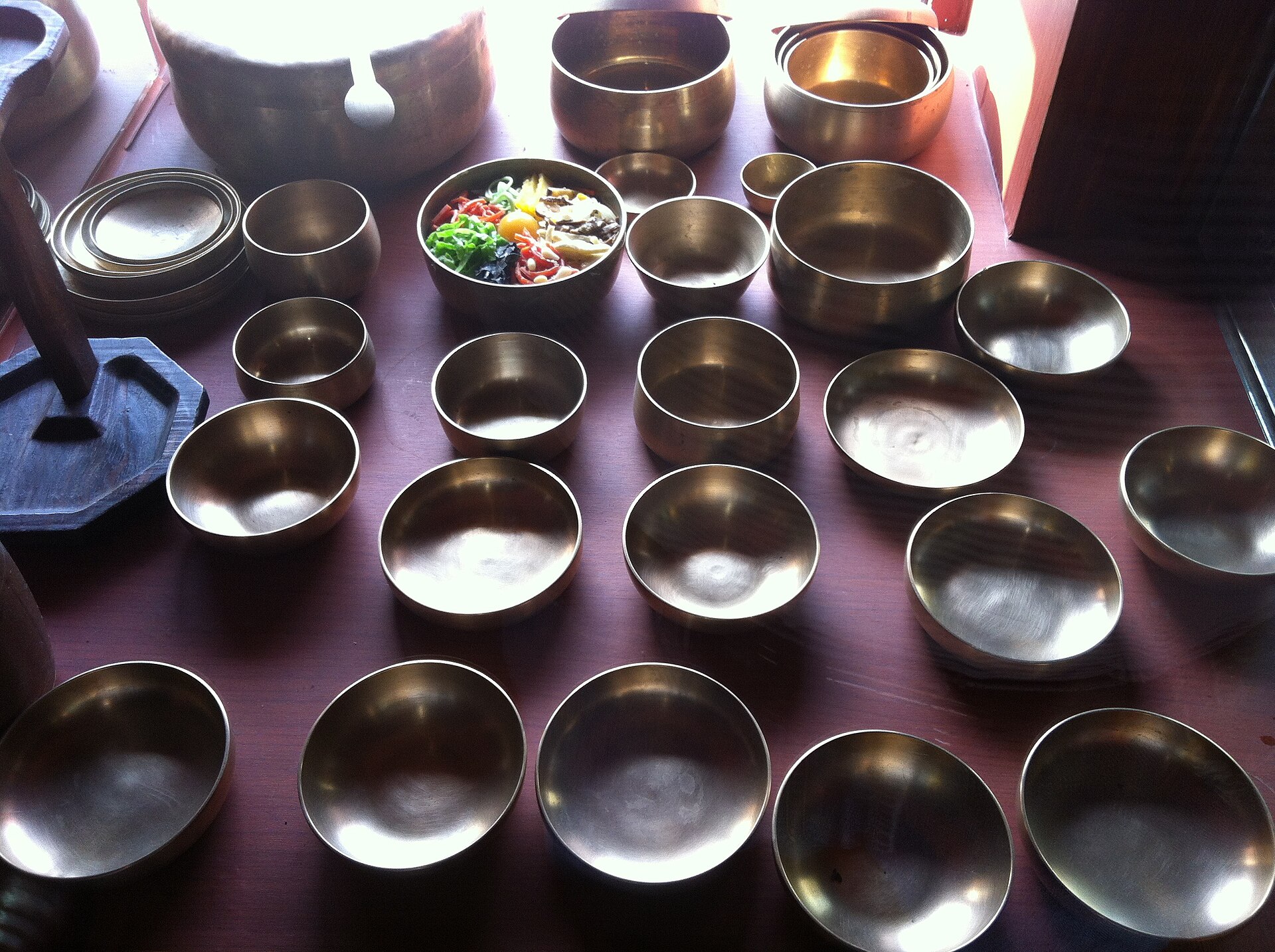 Rows of bangjja yugi bronze bowls of various sizes on a table, one of them filled with bibimbap