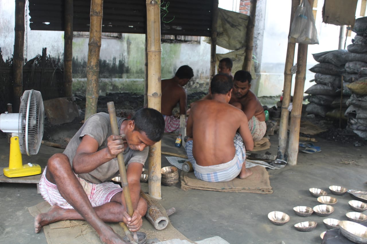 Artisans seated on the floor finishing bell-metal bowls with long-handled tools in a workshop in Sarthebari, Assam, India