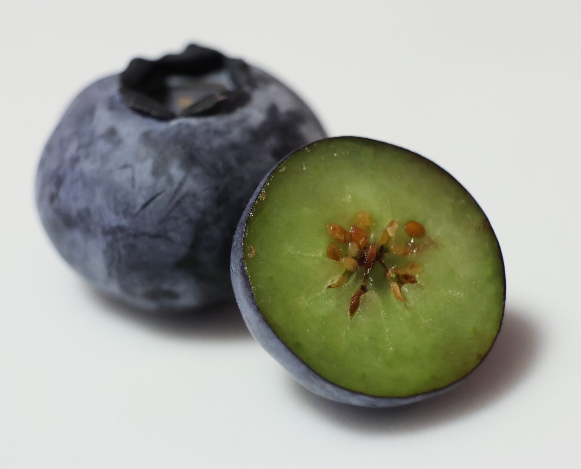 A whole blueberry sits next to one cut in half. The outer skin is deep blue-purple, while the cut flesh inside is pale green
