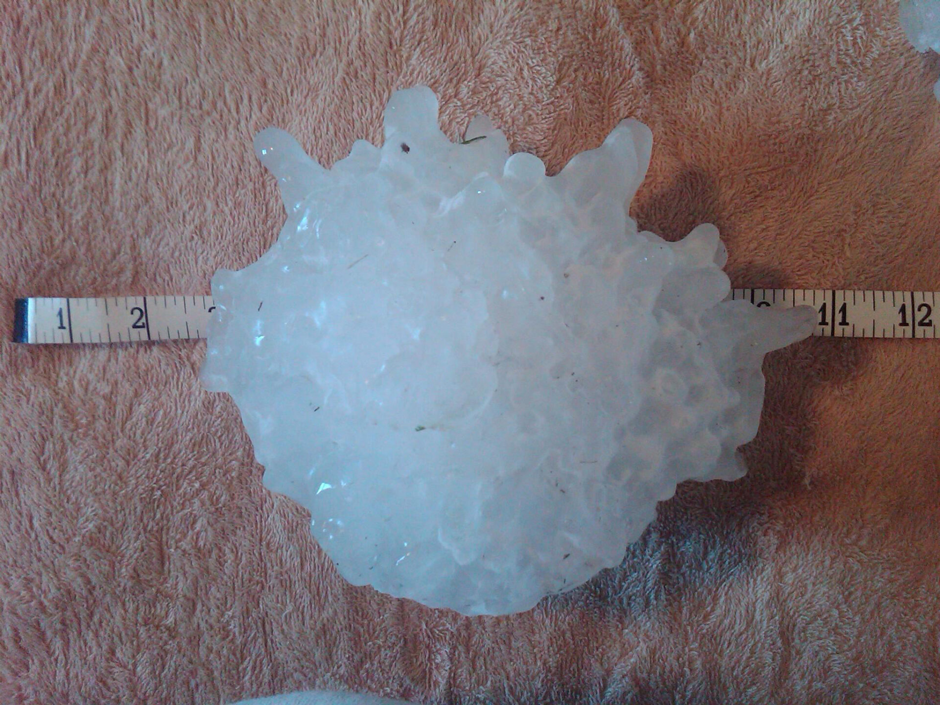 A very large, lumpy hailstone resting on a tape measure, with the marking pointing to around 11-12 cm