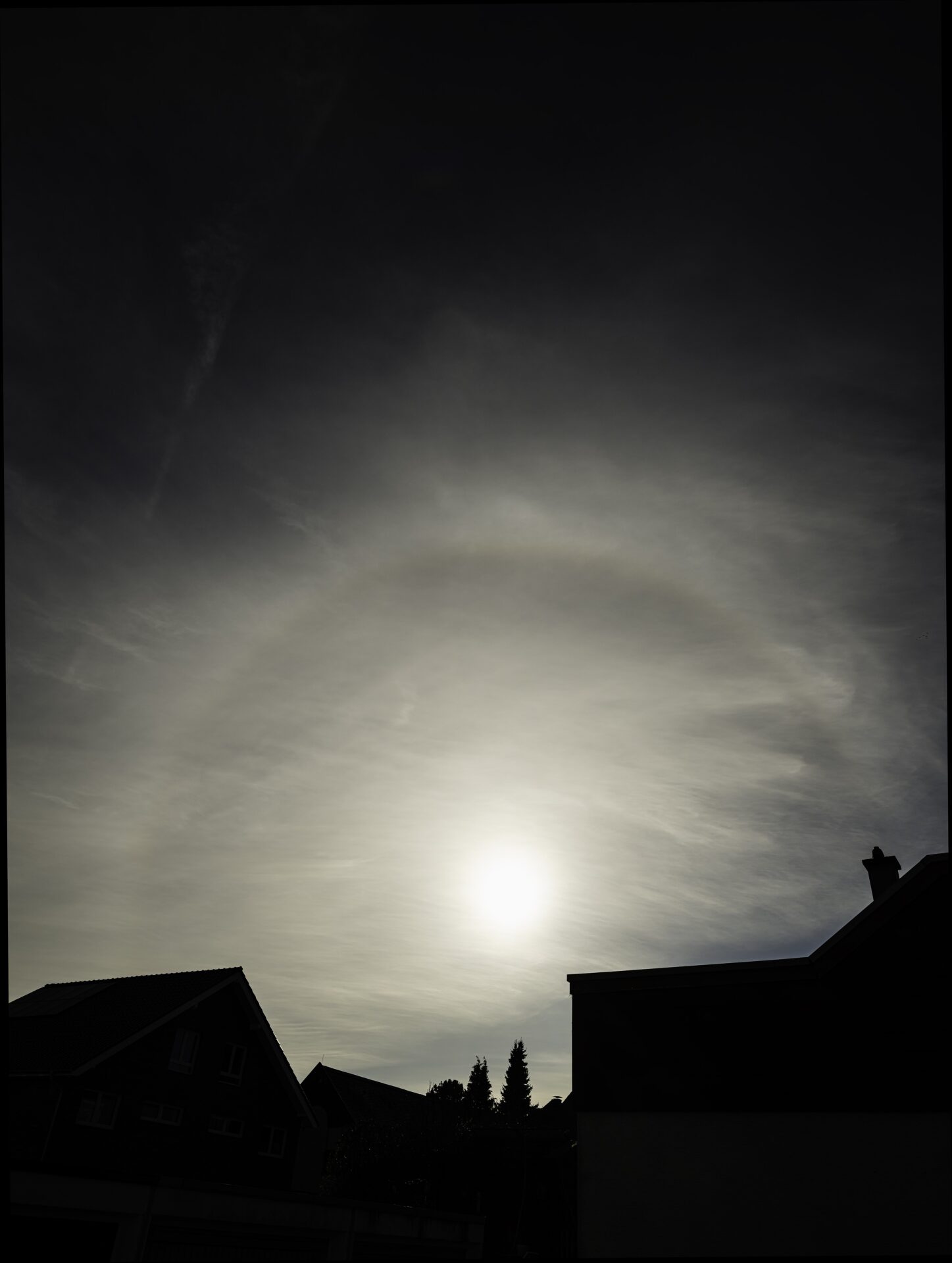 The upper arc of a solar halo in a thin cloud layer above rooftops