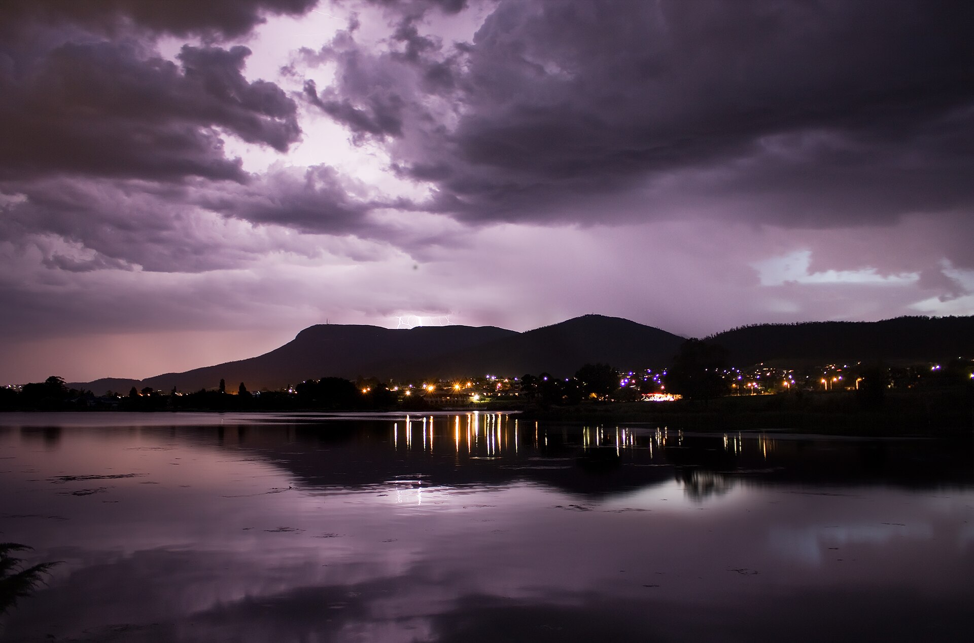 Illuminated clouds and faint lightning above Mount Wellington, with shoreline lights reflected in the water
