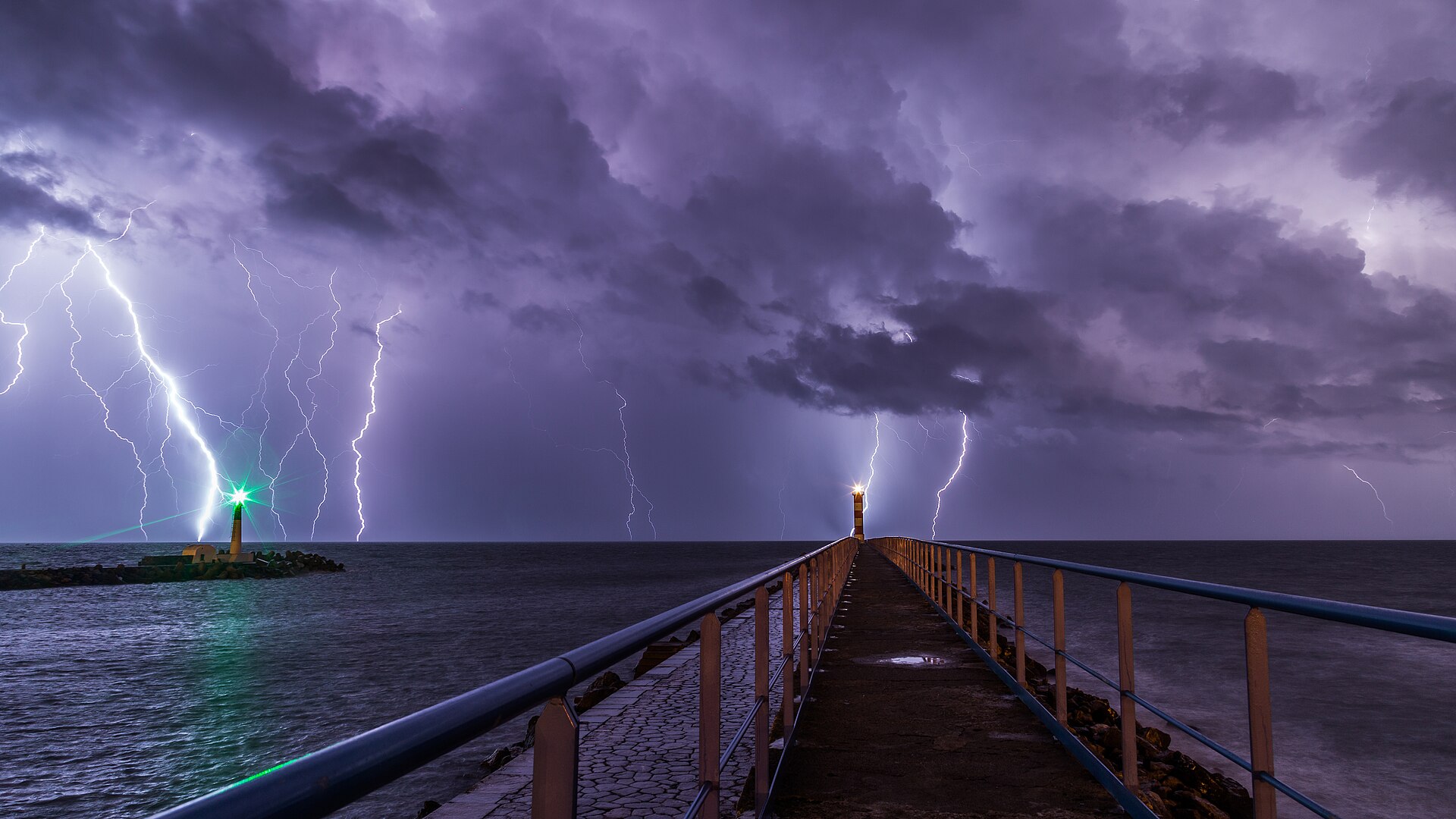 A long-exposure photograph of multiple lightning channels over the sea beyond a breakwater and two lighthouses at Port-la-Nouvelle, France