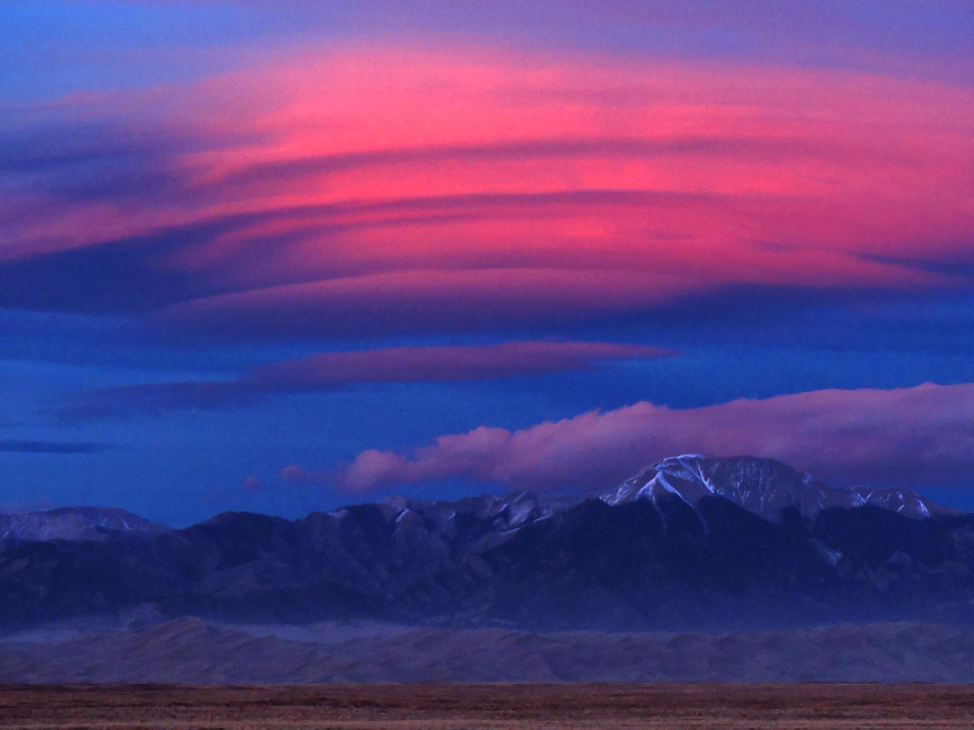 A pink, multilayered lenticular cloud above snow-dusted mountains