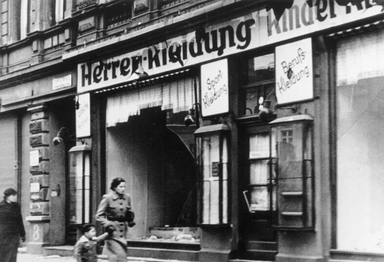 Passers-by walk past a clothing shop whose windows are smashed and whose door stands open, in a black-and-white photo.