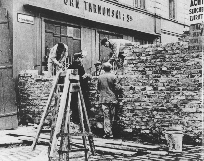 Workers with a ladder laying bricks to build a wall across a street, in a black-and-white photo.