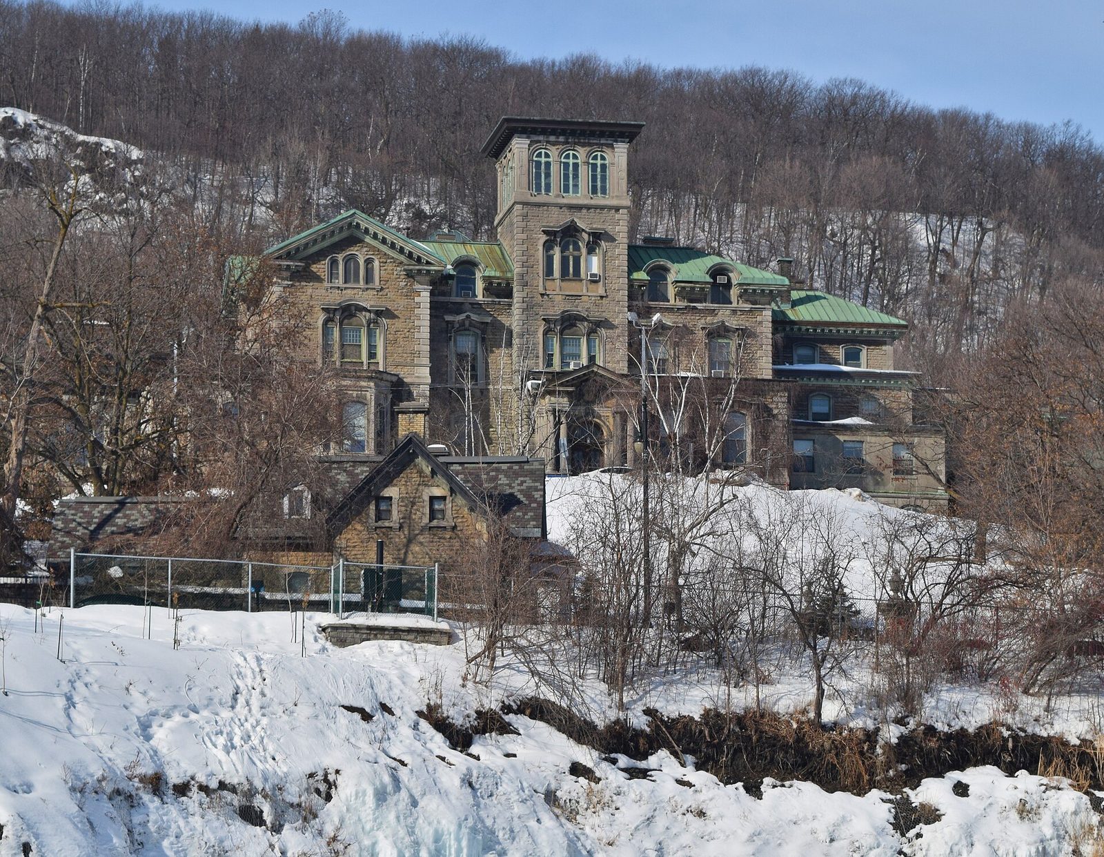 Winter photograph of a towered stone mansion standing below a wooded, snow-covered hillside